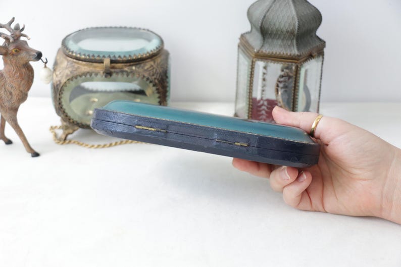 May include: A vintage, rectangular jewelry box with a teal top and dark blue sides. The box is held in a hand, with other antique decorative items in the background, including a deer figurine and a glass-covered container.