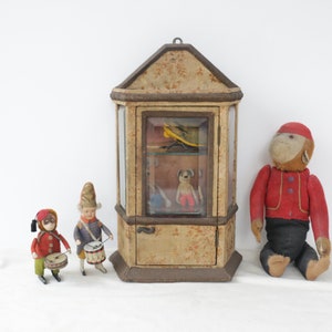 May include: A vintage wooden display case with glass doors, featuring a small stuffed monkey wearing a red jacket, a toy dog, and a toy aeroplane. Two wind-up toy drummers stand in front of the case.