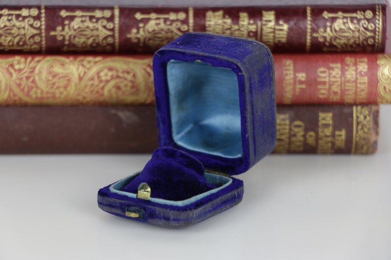May include: An open, antique blue velvet ring box with a light blue satin interior. The box is rectangular with a hinged lid and a small gold-colored clasp. The background features a stack of vintage books.