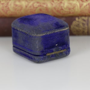 May include: A small, vintage, cobalt blue velvet jewelry box. The box is rectangular with a hinged lid and a gold-colored clasp. The velvet has a slightly worn appearance, suggesting age. The background includes antique books.