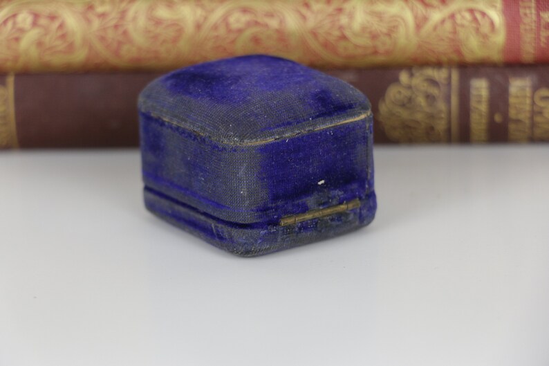 May include: A small, vintage, cobalt blue velvet jewelry box. The box is rectangular with a hinged lid and a gold-colored clasp. The velvet has a slightly worn appearance, suggesting age. The background includes antique books.