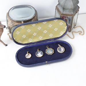 May include: An open, blue velvet jewelry case with four small, gold-framed pendants. The pendants feature miniature portraits of animals and people. A decorative deer figurine and other antique items are in the background.