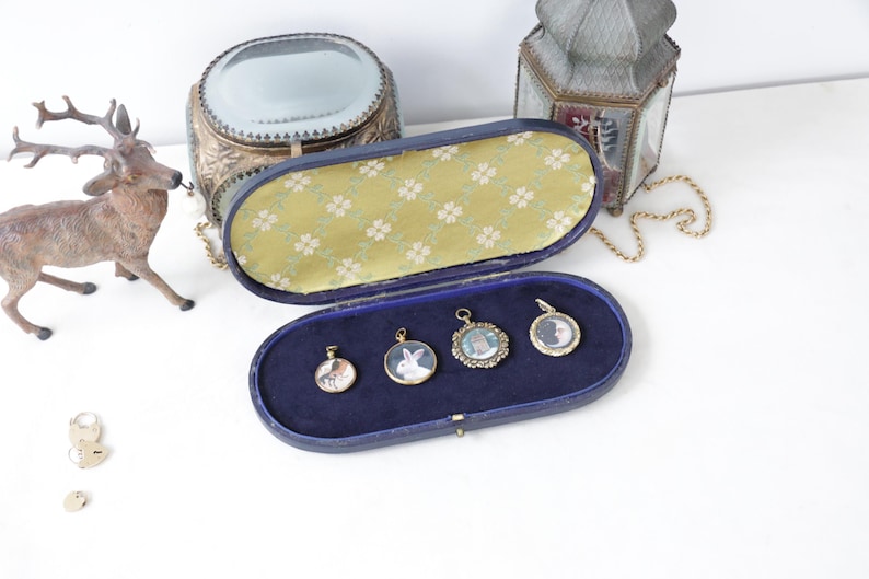 May include: An open, blue velvet jewelry case with four small, gold-framed pendants. The pendants feature miniature portraits of animals and people. A decorative deer figurine and other antique items are in the background.