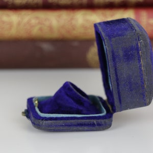 May include: An open, antique blue velvet ring box. The box is rectangular with a hinged lid and a plush interior. The velvet is a deep blue, showing signs of age. The box is likely designed to hold a ring or other small jewelry item.