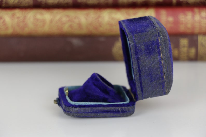May include: An open, antique blue velvet ring box. The box is rectangular with a hinged lid and a plush interior. The velvet is a deep blue, showing signs of age. The box is likely designed to hold a ring or other small jewelry item.