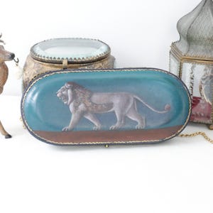 May include: An oval-shaped, teal-colored decorative box with a painted lion illustration. The box has a brown base and a gold-colored trim. Other decorative items include a deer figurine, a gold-colored lantern, and a decorative jar.