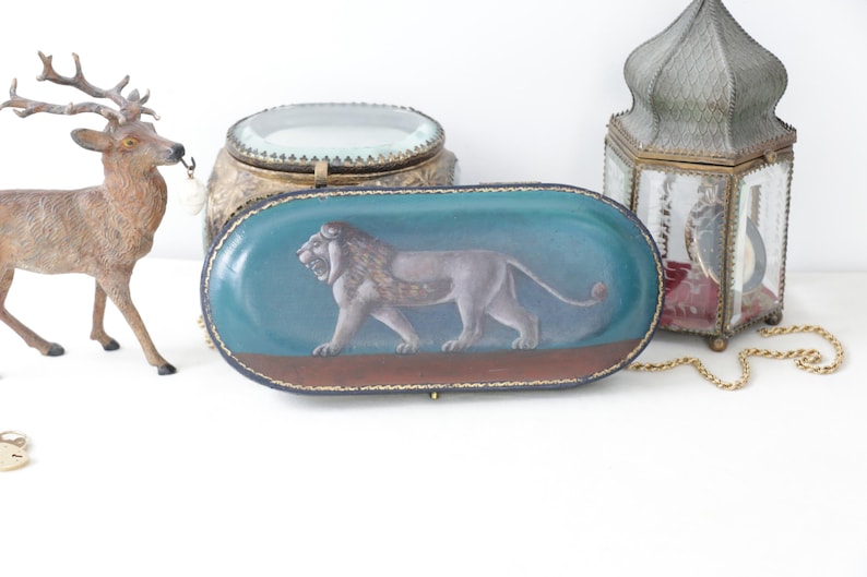 May include: An oval-shaped, teal-colored decorative box with a painted lion illustration. The box has a brown base and a gold-colored trim. Other decorative items include a deer figurine, a gold-colored lantern, and a decorative jar.