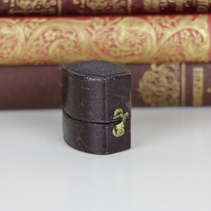 May include: A small, brown leather ring box with a brass clasp. The box is closed and sitting on a white surface.