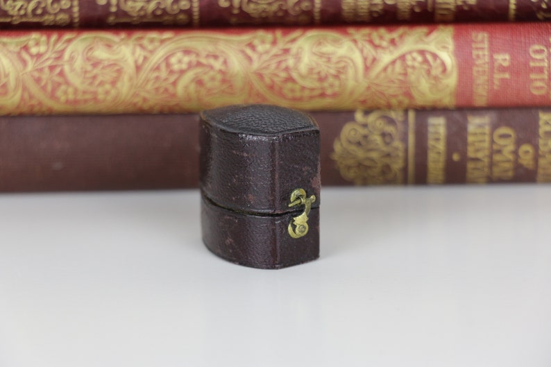 May include: A small, brown leather ring box with a brass clasp. The box is closed and sitting on a white surface.