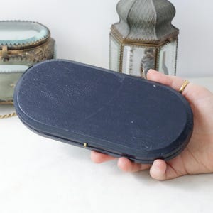 May include: A navy blue, oval-shaped box with a textured surface. The box is held in a hand, with other decorative items in the background. The box has a small, gold-colored clasp.