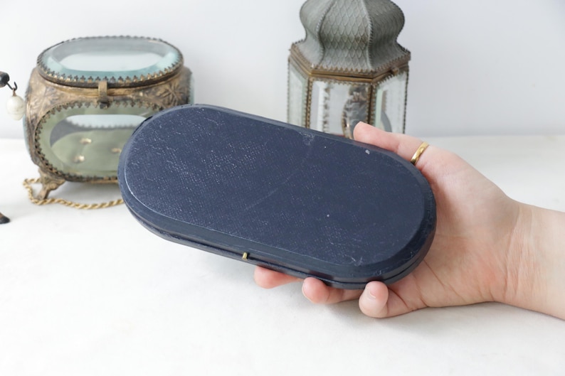 May include: A navy blue, oval-shaped box with a textured surface. The box is held in a hand, with other decorative items in the background. The box has a small, gold-colored clasp.
