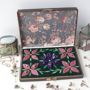 May include: A vintage jewellery box with a floral interior and a dark green velvet base. The base features embroidered pink and purple flower designs. A small, ornate jewellery box and a decorative lantern are also present.