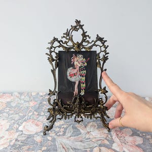 Könnte beinhalten: Verzierter bronzefarbener Bilderrahmen mit schwarzem Hintergrund, der eine farbenfrohe Illustration von zwei kostümierten Figuren zeigt. Der Rahmen hat ein aufwendiges, scrollendes Design und steht auf einer floral gemusterten Oberfläche.