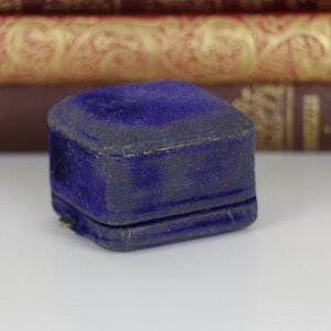 May include: A small, vintage, square jewelry box. The box is covered in a deep blue velvet fabric, showing signs of wear. The box is closed, with a visible seam. Background includes antique books.