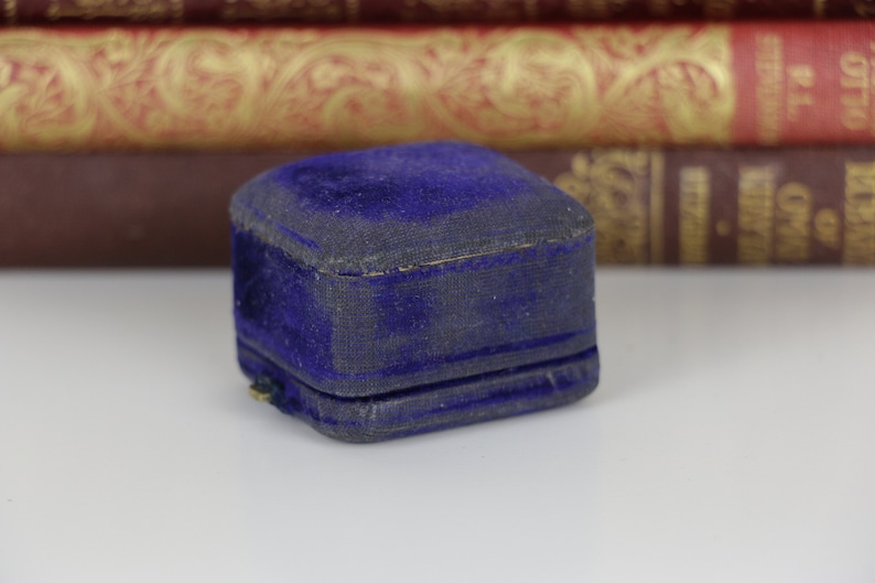May include: A small, vintage, square jewelry box. The box is covered in a deep blue velvet fabric, showing signs of wear. The box is closed, with a visible seam. Background includes antique books.