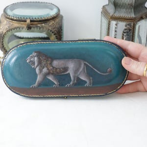 May include: An oval-shaped, teal-colored box with a detailed lion illustration. The lion is depicted in shades of gray and brown, set against a teal background. The box has a brown base and gold trim, suggesting a vintage or antique style.