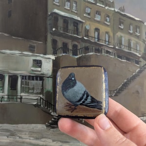 May include: A small square wooden plaque featuring a hand-painted grey and black pigeon on a tan background. The edges of the plaque are painted dark blue with gold accents. The pigeon is depicted in profile, facing left, with detailed feathers.