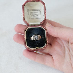 May include: A gold ring featuring a miniature skull image within a square frame, presented in an open, red leatherette jewellery box. The box has a black velvet interior and a printed label on the lid.