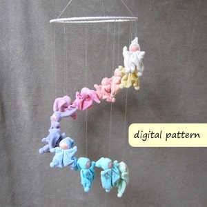 May include: A rainbow-colored mobile with felt figures hanging from a white metal ring. The figures are in various shades of pink, purple, blue, and green. The mobile is labeled "digital pattern EN".