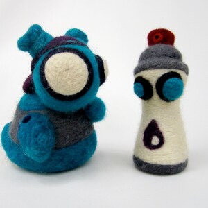 May include: Two felt toys, one is a blue and gray creature with a white face and the other is a gray and white spray paint can with blue eyes.