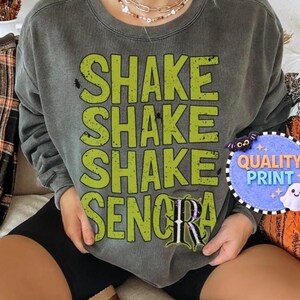 May include: A charcoal gray crewneck sweatshirt featuring the lime green text SHAKE SHAKE SHAKE SENORA printed in a distressed font. The letter R in the final word is stylized with a purple and black pattern, and small black spider icons are visible.