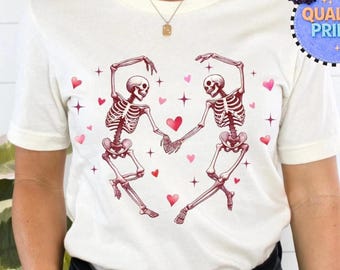 May include: A white short-sleeve t-shirt featuring a graphic of two skeletons dancing and holding hands to form a heart shape. The design includes small red hearts and four-pointed stars scattered around the skeletons on the front of the shirt.