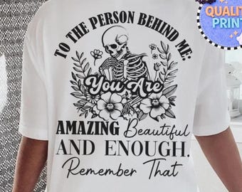 May include: A white t-shirt featuring a black graphic on the back. The design includes a skeleton surrounded by flowers and the text: TO THE PERSON BEHIND ME: YOU ARE AMAZING BEAUTIFUL AND ENOUGH. REMEMBER THAT. The print is centered on the shirt.