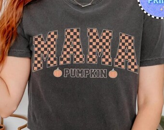 May include: A charcoal gray short-sleeve t-shirt featuring the word MAMA in a large, orange and black checkered block font. Below the word, the text PUMPKIN is printed in a simple font, flanked by two small orange pumpkin illustrations.