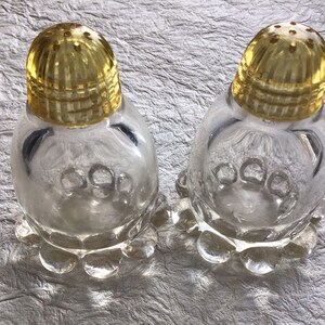 May include: Two clear glass salt and pepper shakers with yellow tops. The shakers have a scalloped base and a textured design.