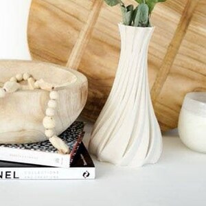 May include: A white, textured vase holds green eucalyptus branches, set against a wooden cutting board and a wooden bowl with beads. A stack of books with the word "CHANEL" is visible.