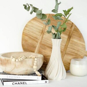 May include: A still life arrangement featuring a wooden cutting board, a white vase with eucalyptus, a wooden bowl with beads, a candle in a glass holder, and two books. The items are arranged on a white surface.