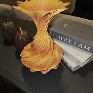 May include: A decorative, abstract vase with a sunflower-like top and a swirling, layered design. The vase is primarily yellow and orange, with a flared top and a narrow base. Two small decorative pumpkins are in the background.