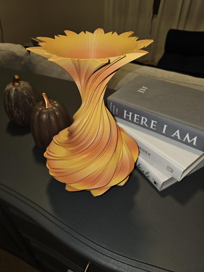 May include: A decorative, abstract vase with a sunflower-like top and a swirling, layered design. The vase is primarily yellow and orange, with a flared top and a narrow base. Two small decorative pumpkins are in the background.