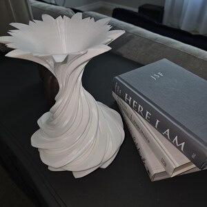 May include: A white, abstract decorative vase with a flower-like top and a twisted, swirling base. The vase is next to a stack of books with the title "HERE I AM" visible.