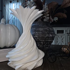 May include: A white, sculptural vase with a swirling design is the focal point of this still life. A white pumpkin and a dark, textured vase with dried floral arrangements are also present. A decorative black metal bicycle adds a touch of whimsy to the scene.