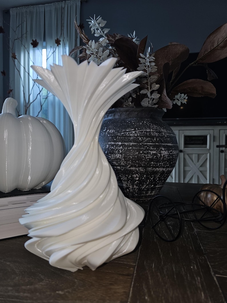 May include: A white, sculptural vase with a swirling design is the focal point of this still life. A white pumpkin and a dark, textured vase with dried floral arrangements are also present. A decorative black metal bicycle adds a touch of whimsy to the scene.