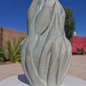 Könnte beinhalten: Eine hellblaue Keramikskulptur mit einem strukturierten, fließenden Design. Die abstrakte Form hat eine glänzende Oberfläche und wird auf einer weißen Oberfläche präsentiert. Die Skulptur ähnelt drapiertem Stoff oder einer stilisierten organischen Form.