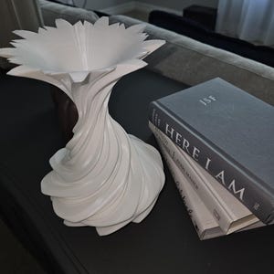 May include: A white, abstract decorative vase with a flower-like top and a twisted, swirling base. The vase is placed on a dark surface next to a stack of books with the title "HERE I AM".