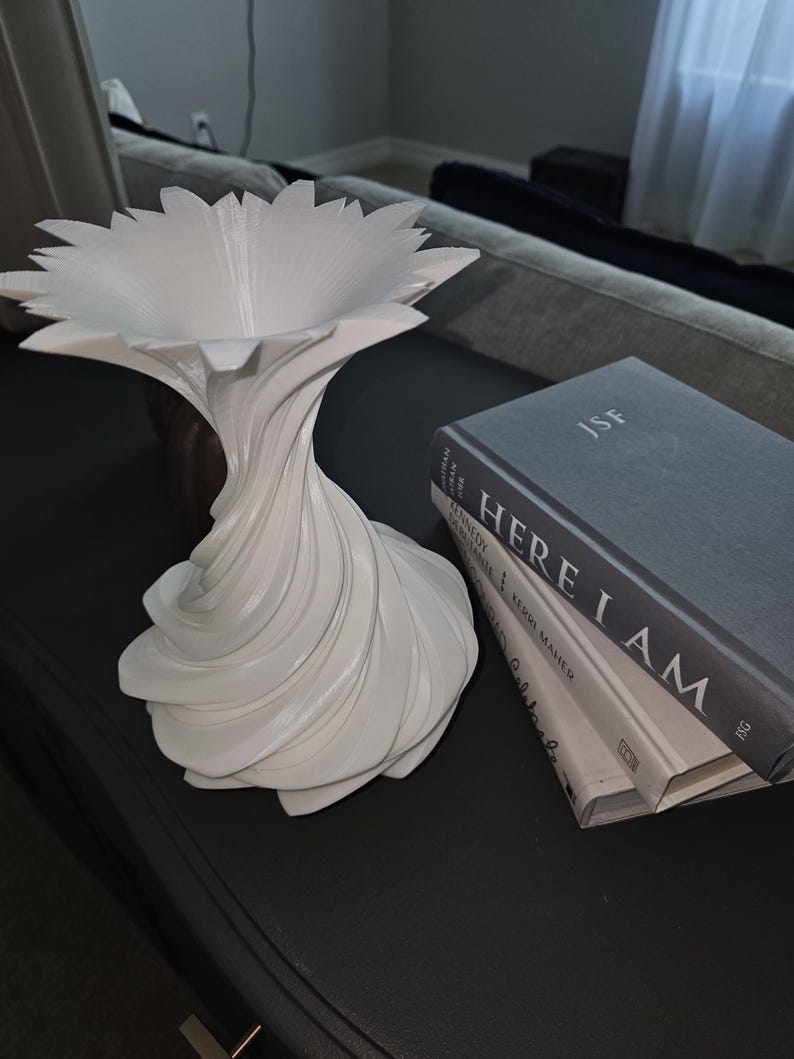 May include: A white, abstract decorative vase with a flower-like top and a twisted, swirling base. The vase is placed on a dark surface next to a stack of books with the title "HERE I AM".