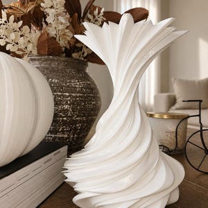 May include: A white, sculptural vase with a unique, swirling design. The vase has a flared top and a textured, layered appearance. It is displayed on a wooden surface, alongside other decorative items.
