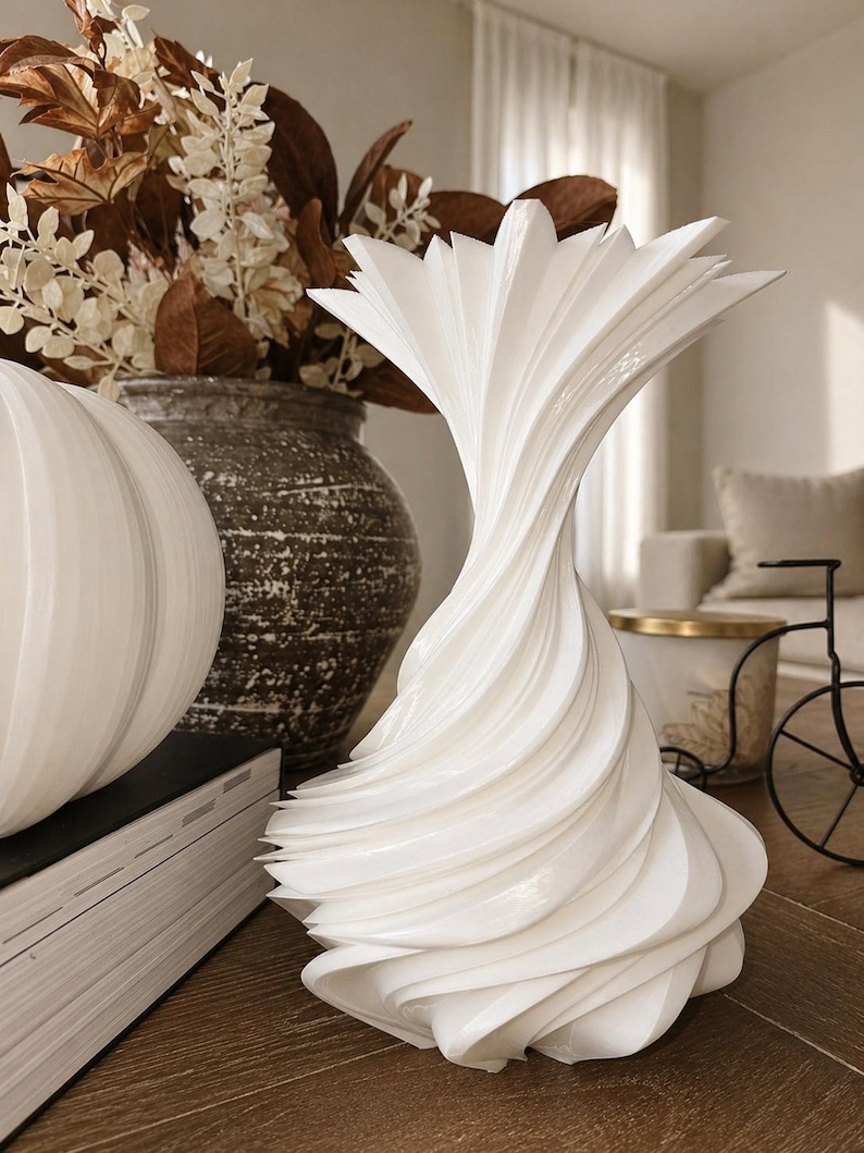 May include: A white, sculptural vase with a unique, swirling design. The vase has a flared top and a textured, layered appearance. It is displayed on a wooden surface, alongside other decorative items.
