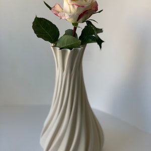 May include: A cream-colored, spiral-shaped vase holds a single rose. The rose has white petals edged with pink and green leaves. The vase has a textured, ridged design and is set against a white background.