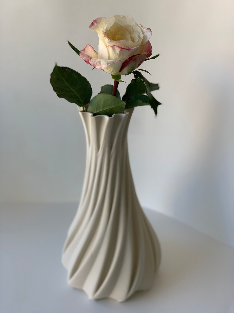 May include: A cream-colored, spiral-shaped vase holds a single rose. The rose has white petals edged with pink and green leaves. The vase has a textured, ridged design and is set against a white background.
