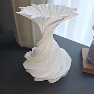 May include: A white, decorative vase with a unique, swirling design. The vase has a flared, flower-like top and a textured, layered base. The overall shape is reminiscent of a blooming flower. The vase is made of a solid, opaque material.