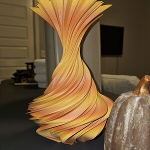 May include: A decorative, abstract vase with a unique, layered design. The vase features a spiral shape with alternating layers of yellow and orange hues. The vase is a modern home decor item.