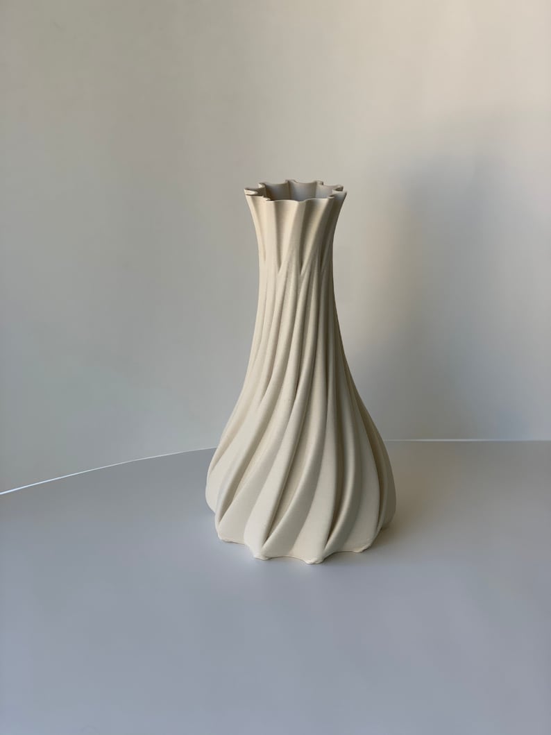May include: A tall, cream-colored vase with a unique, swirling design. The vase has a ruffled top and a wide base, tapering upwards to a narrower neck. The textured surface creates a dynamic, eye-catching aesthetic. The vase is suitable for home decor.