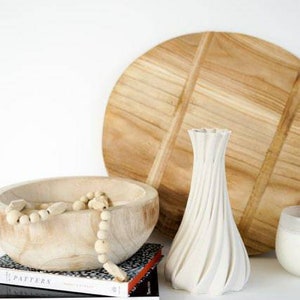 May include: A white, textured vase with a unique spiral design stands next to a wooden bowl filled with decorative beads. A large wooden cutting board and books with the word "CHANEL" are also visible.