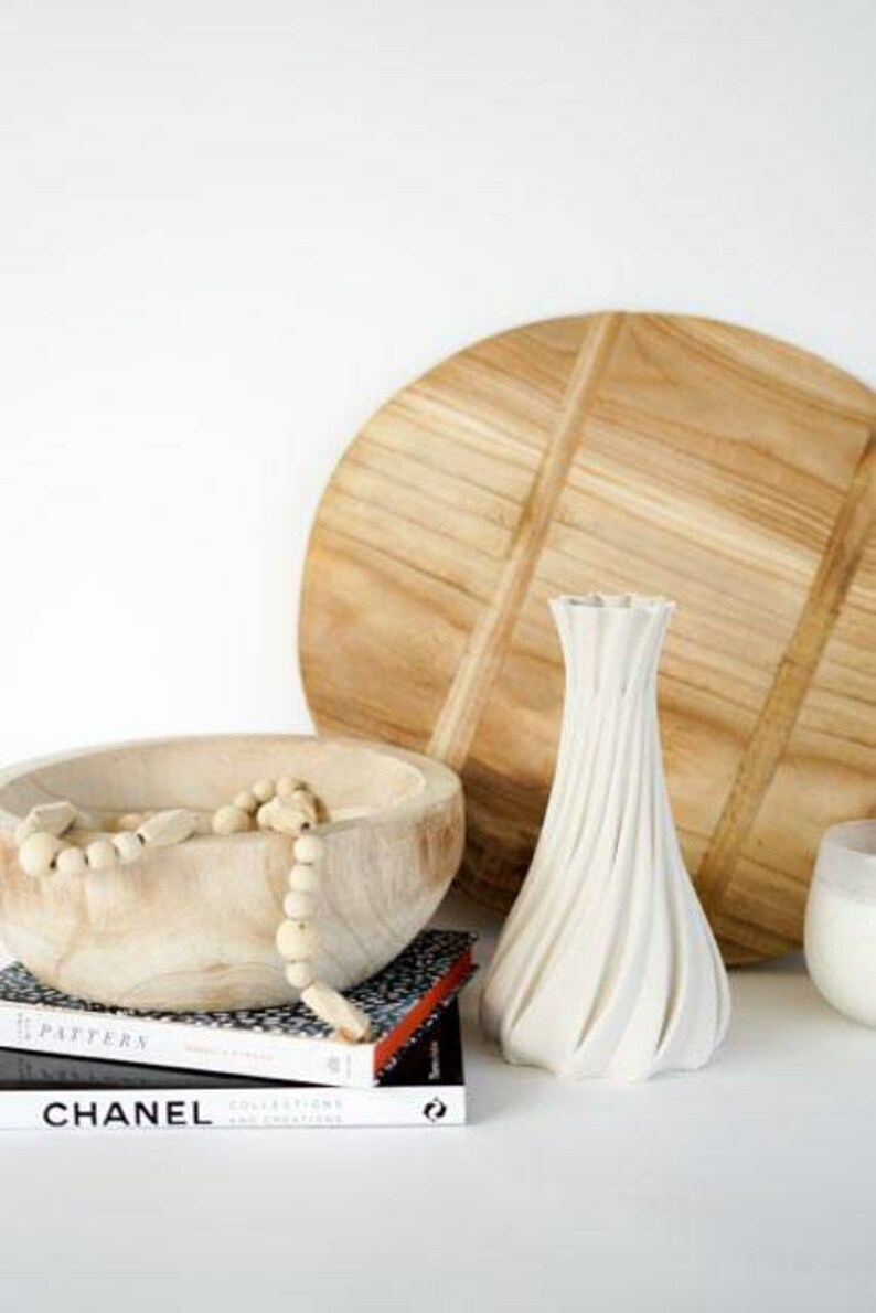 May include: A white, textured vase with a unique spiral design stands next to a wooden bowl filled with decorative beads. A large wooden cutting board and books with the word "CHANEL" are also visible.