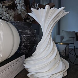 May include: A white, sculptural vase with a spiral design. The vase has a flared top and a textured, layered appearance. Other decorative items, including a white pumpkin and a dark vase with dried flowers, are in the background.