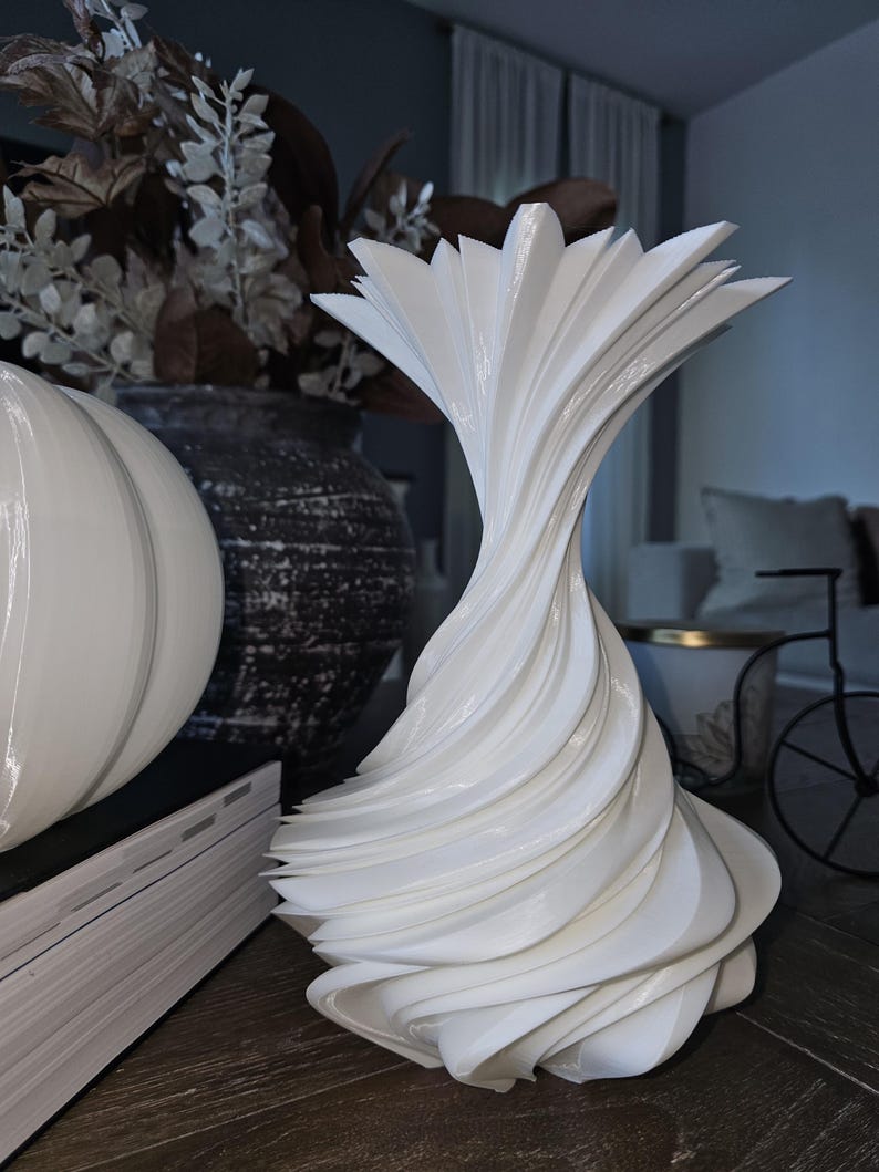 May include: A white, sculptural vase with a spiral design. The vase has a flared top and a textured, layered appearance. Other decorative items, including a white pumpkin and a dark vase with dried flowers, are in the background.